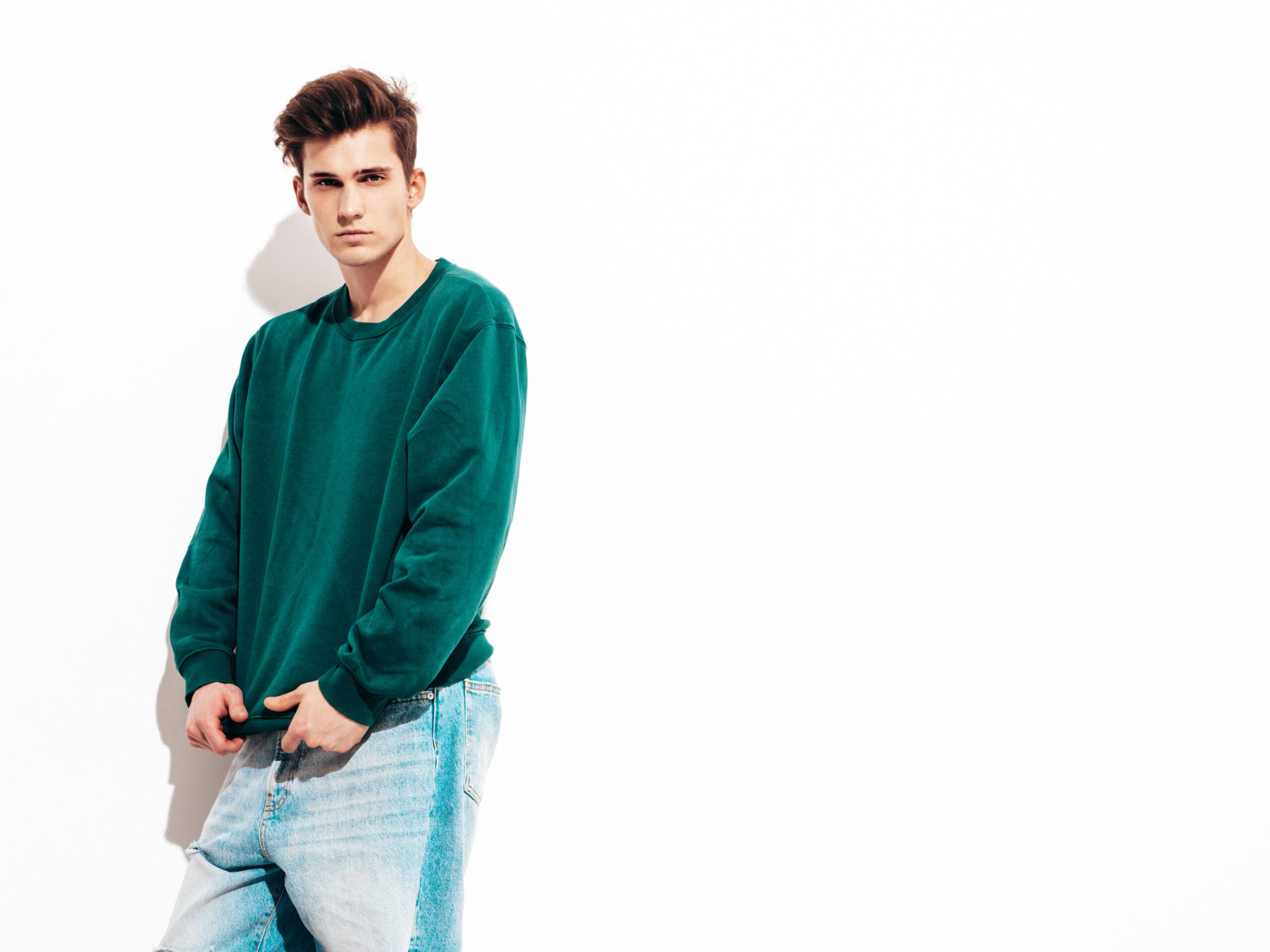 Portrait of handsome confident stylish hipster lambersexual model.Man dressed in green sweater and jeans. Fashion male posing in studio near white wall