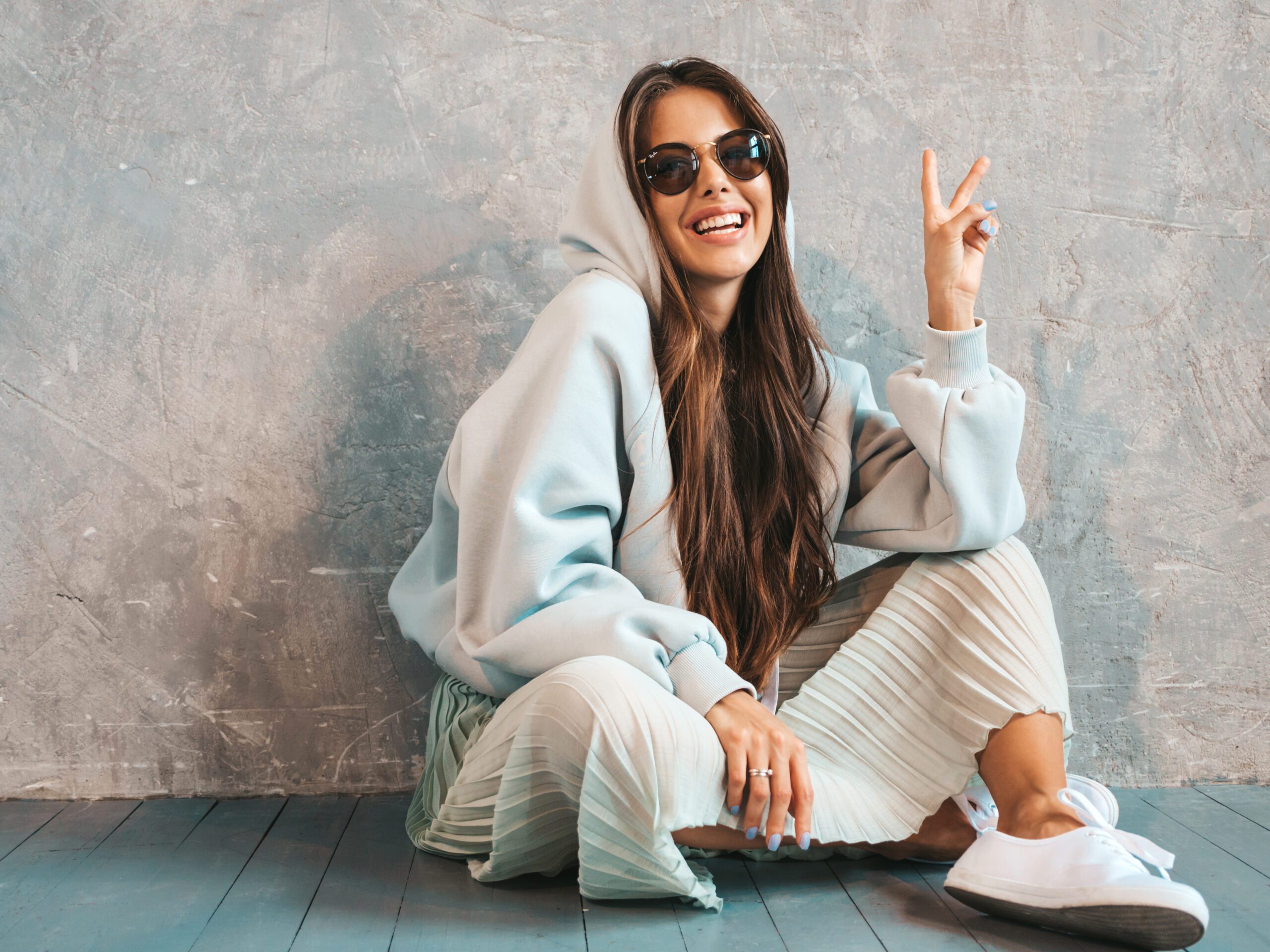 Young beautiful smiling woman shows peace sign. Trendy girl in casual summer hoodie and skirt clothes. Funny and positive female posing near gray wall in studio. In sunglasses sitting on the floor